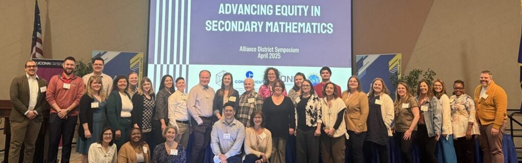 A photo of participants of the Alliance District Symposium lined up for a photo in front a projected slide that says "Advancing Equity in Secondary Mathematics."