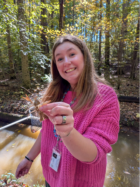 A smiling photo of Alexa Epstein in a creek in the woods. She is wearing a pink shirt and holding a crayfish. She has medium length brown hair.