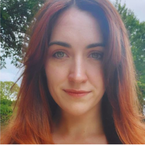 A photo of Caitlin Donovan. She is outside, in front of a tree and is smiling. She has long red hair.