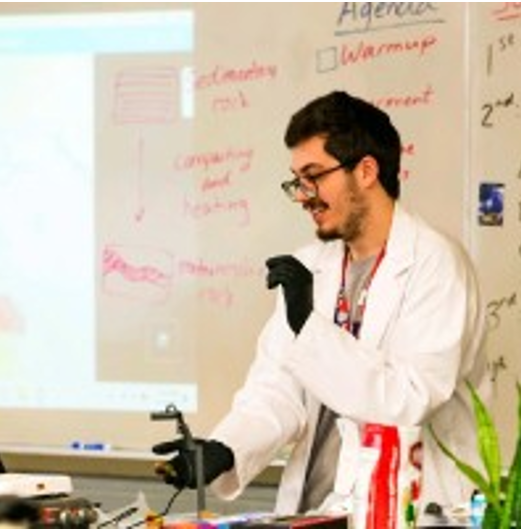 A photo of Colin Dail in a classroom in a white coat. He is wearing glasses and a pair of black gloves and is teaching a lesson.