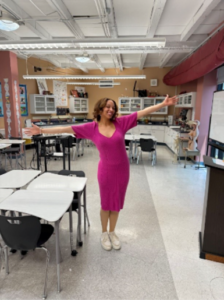 A photo of a person in a pink dress in a classroom with their arms outstretched.